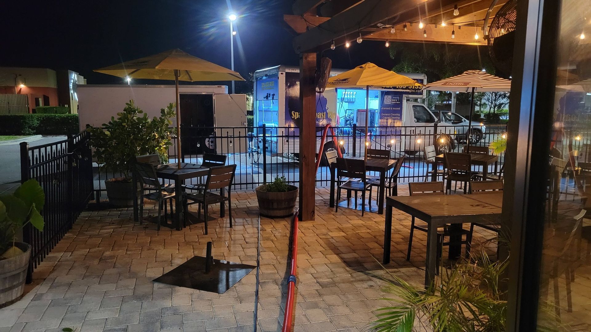 Outdoor restaurant patio at night with tables, chairs, string lights, and food trucks in the background.