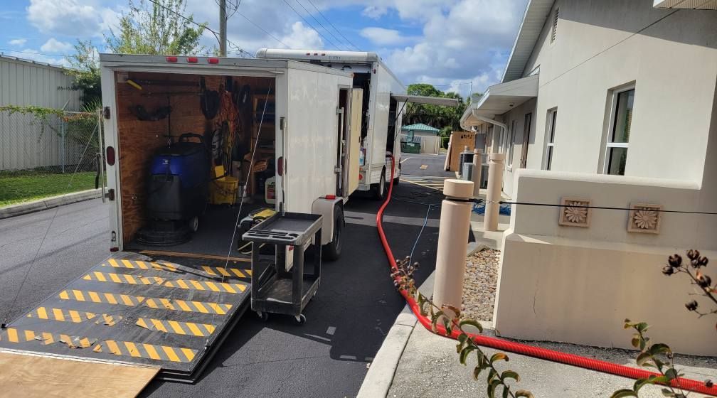 Cargo trailer with ramp open next to a building. Tools and a work cart are visible. A red hose runs nearby.