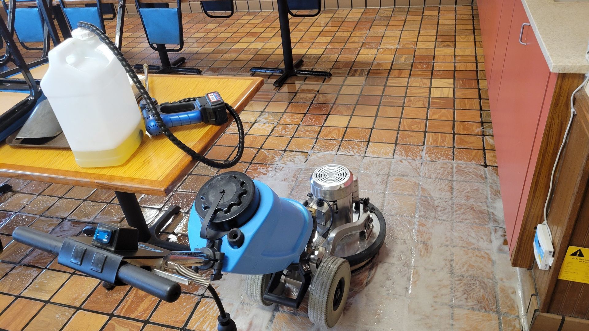 Floor being polished with a machine inside a commercial space with tables, chairs, and a liquid container.