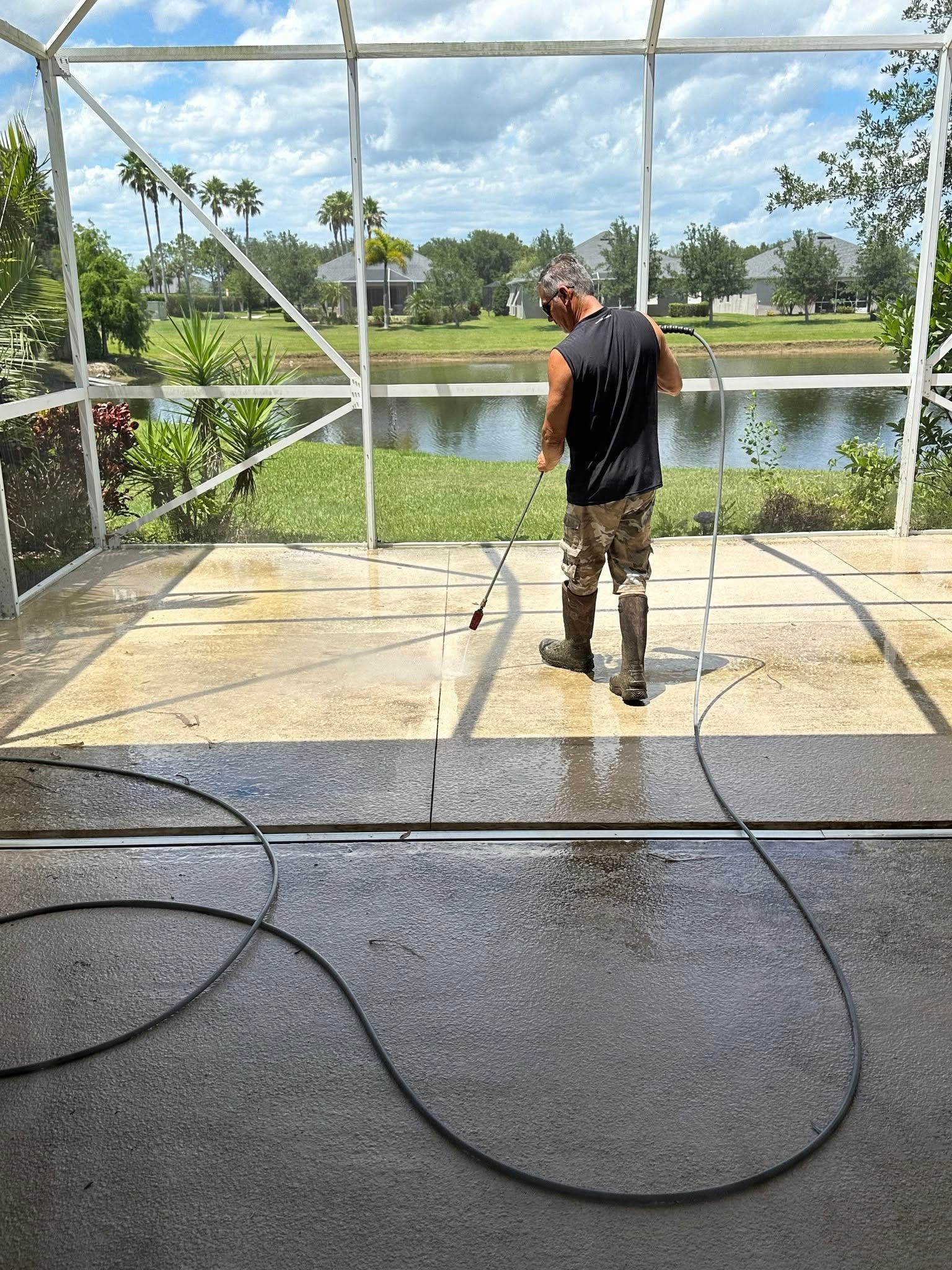 Man pressure washing a concrete patio near a lake under a screened enclosure.