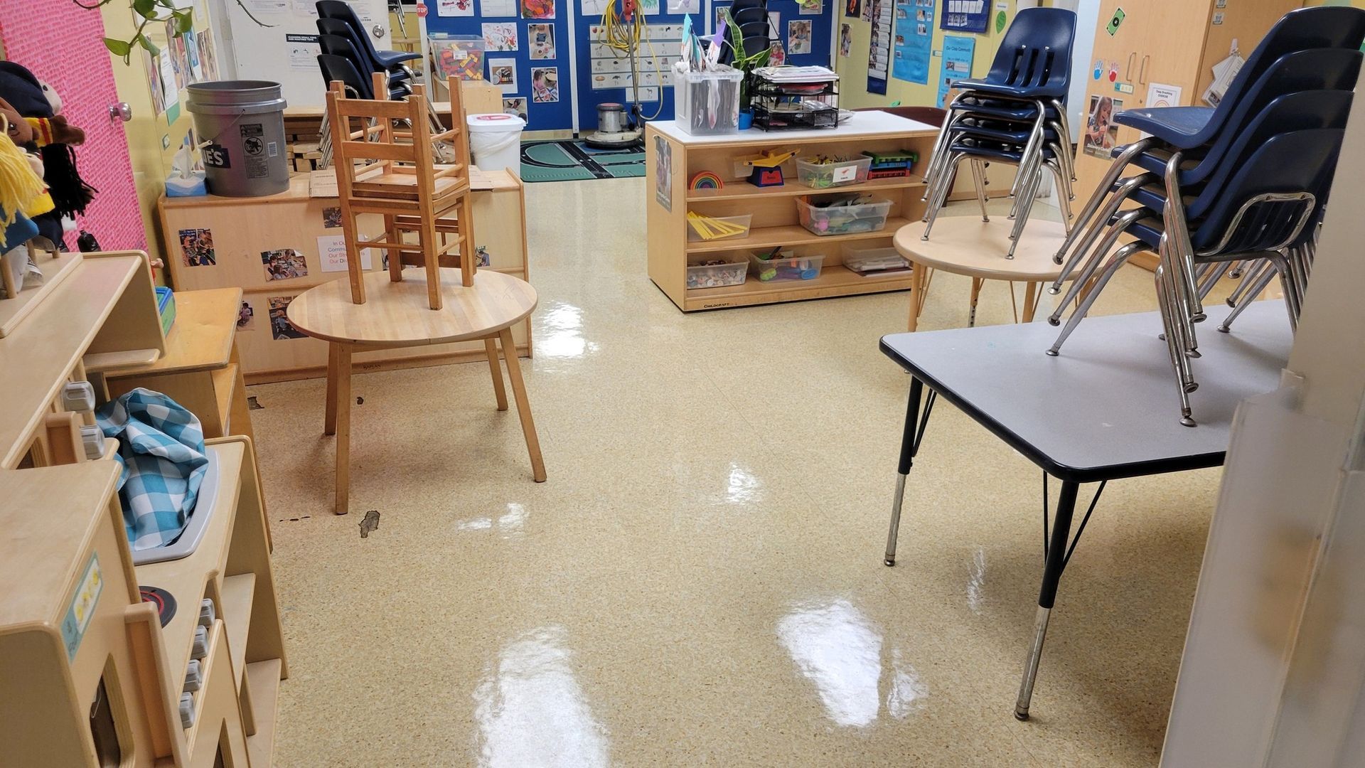 A classroom interior with tables, chairs, and shelves. The floor has a reflective, light-colored surface.
