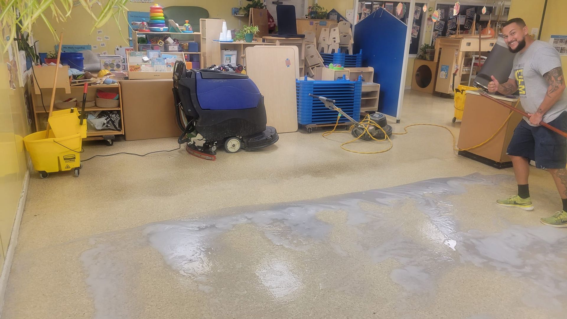 A man spraying water on a floor, cleaning. A floor cleaning machine and yellow bucket are nearby in a room.