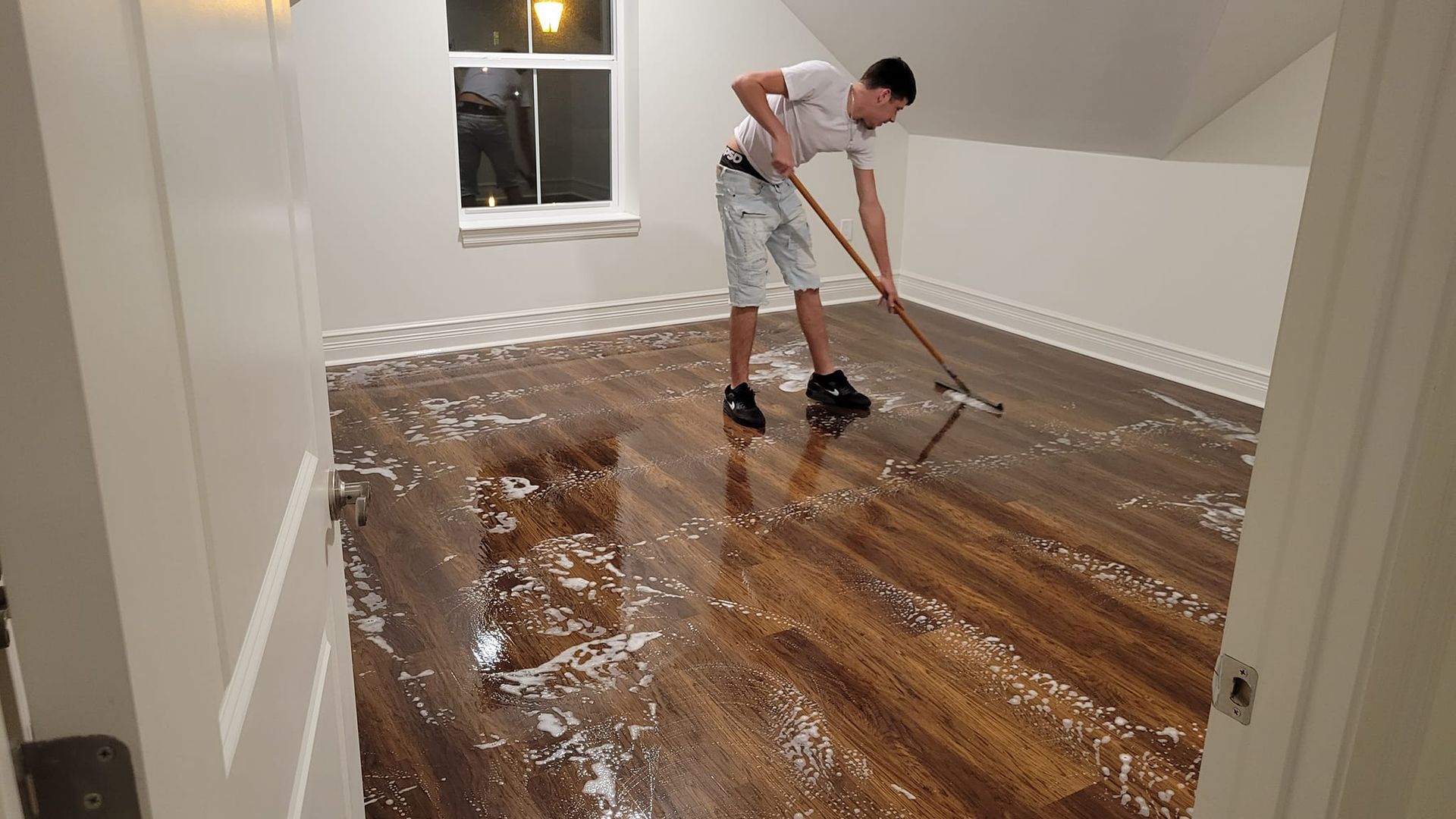A person mopping a wet, wooden floor in a room with white walls and a window.