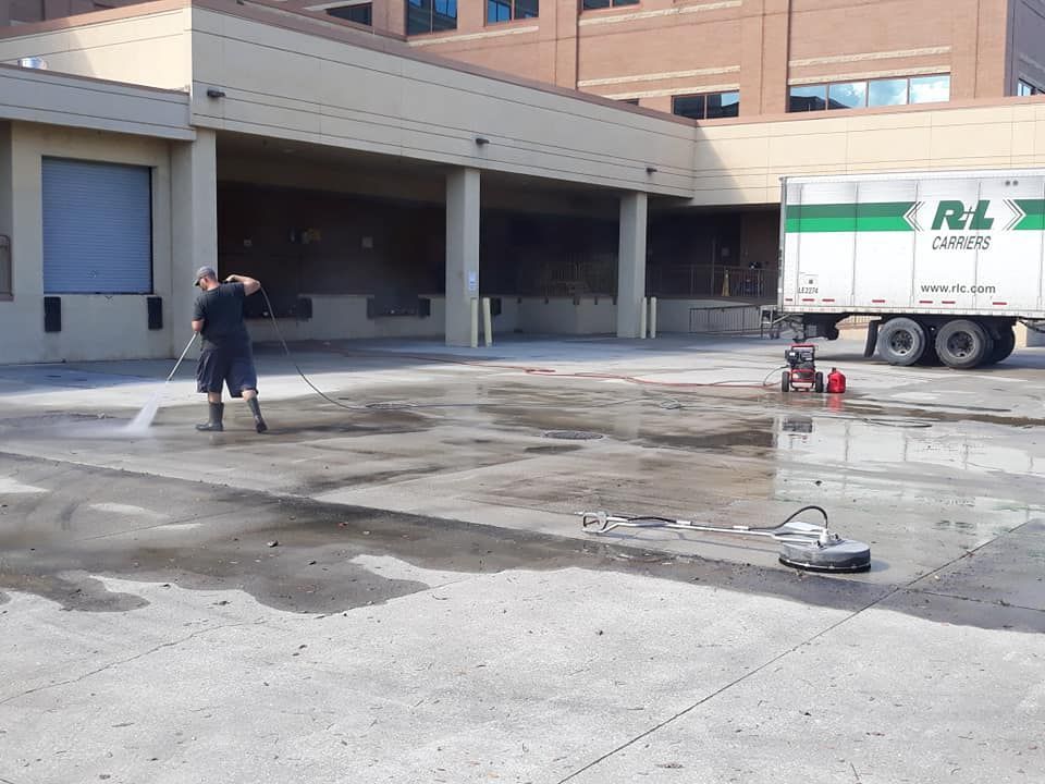 Person pressure washing a concrete loading dock, next to a semi-truck with a green and white trailer.