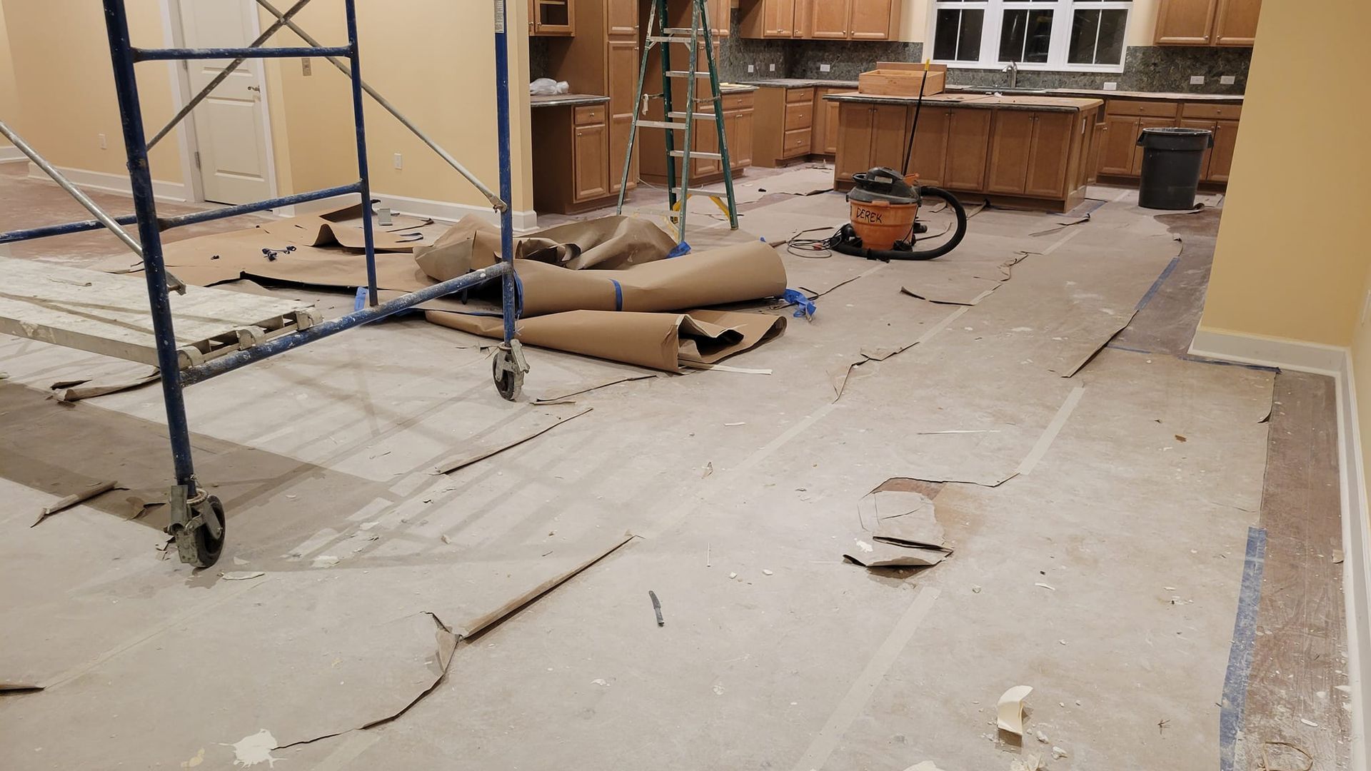 Room undergoing renovation, showing floor demolition, scaffolding, cabinets, and vacuum.