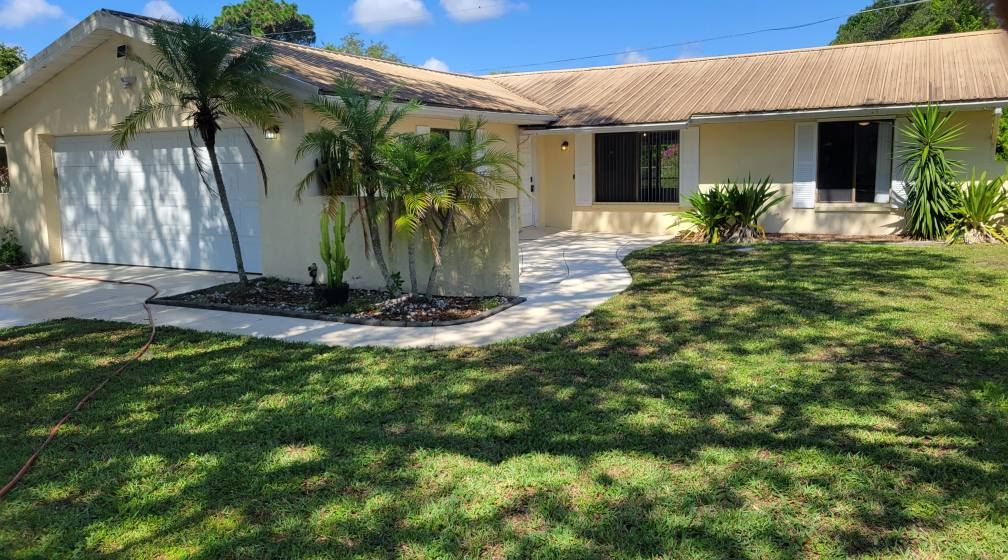 A one-story beige house with a white garage door, palm trees, and a green lawn.