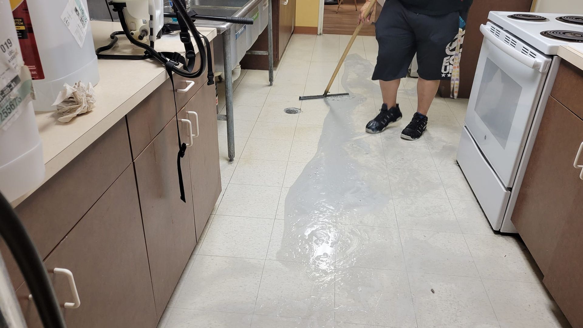Person squeegeeing water off a kitchen floor with brown cabinets and white appliances.