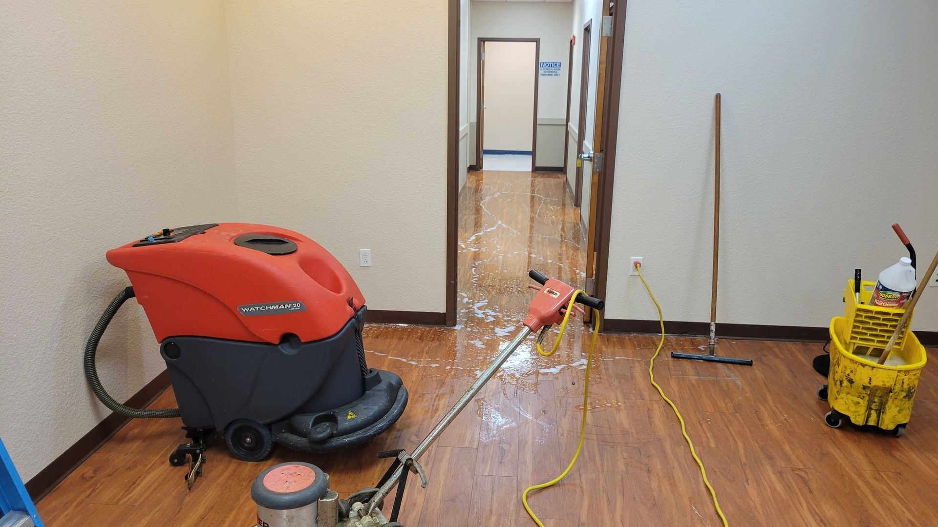 Floor cleaning equipment in a room with wood flooring. A wet floor cleaner, mop, and bucket are present.