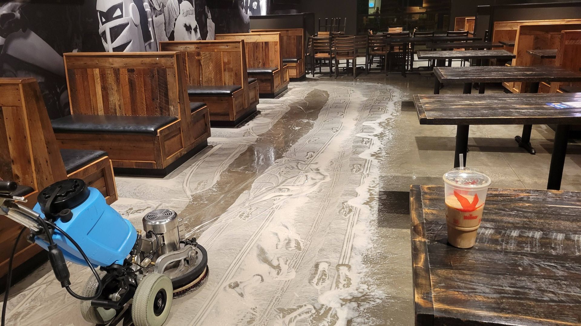 A floor cleaning machine in a coffee shop, cleaning the floor. Brown wooden tables and booths are visible.