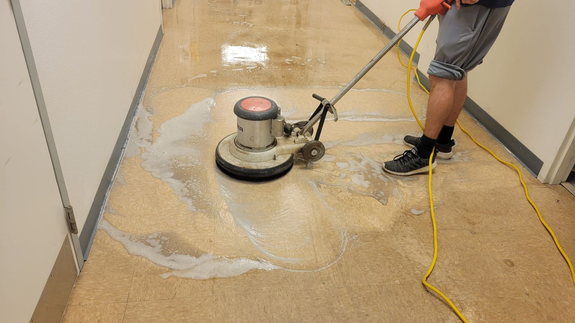 Person using a floor buffer to clean a hallway. Machine sprays cleaning solution.