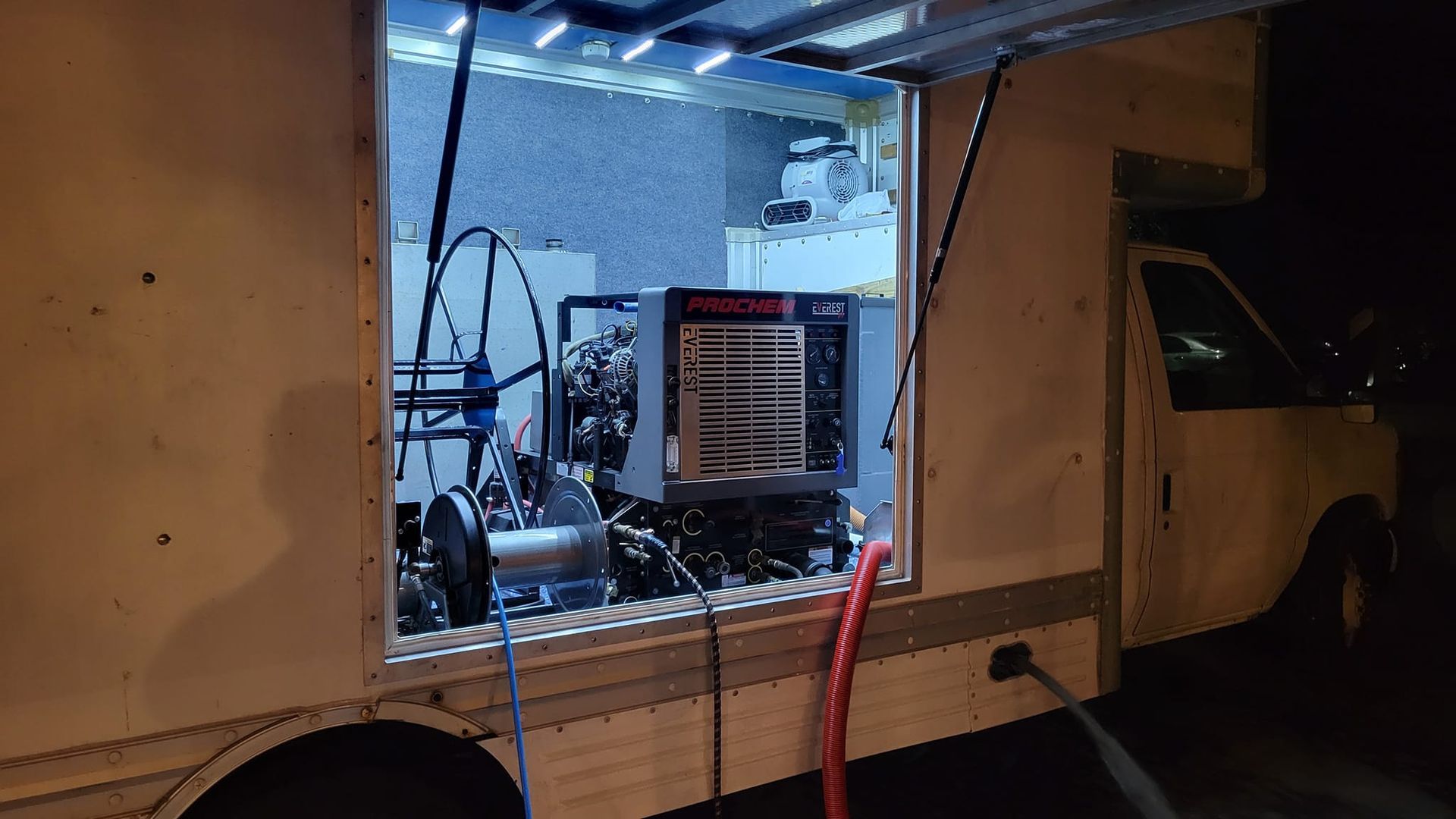 Truck interior with equipment; lit by blue light. Open door reveals machinery and a digital display panel.
