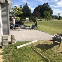 A backyard patio at dusk features a fire pit, low walls with lights, and a stone house backdrop.