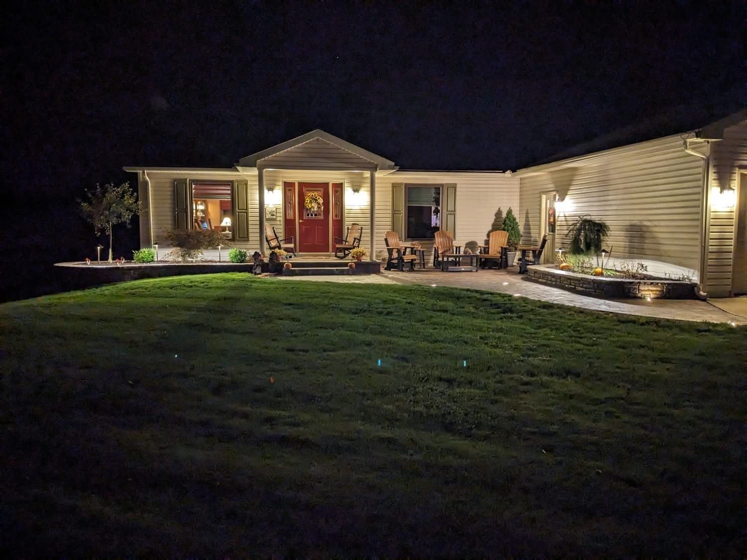 A house at night with a lit front porch and lawn. Red door, sidelights, white siding, and landscape lighting.