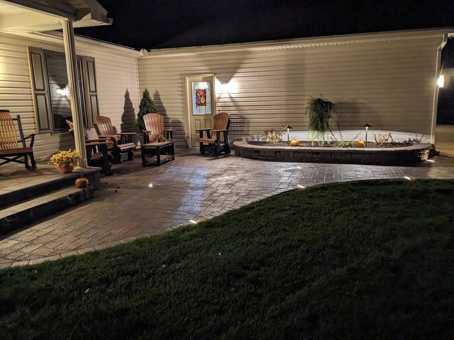 Night view of a brick patio with seating and a small garden, lit by warm lights. House in the background.