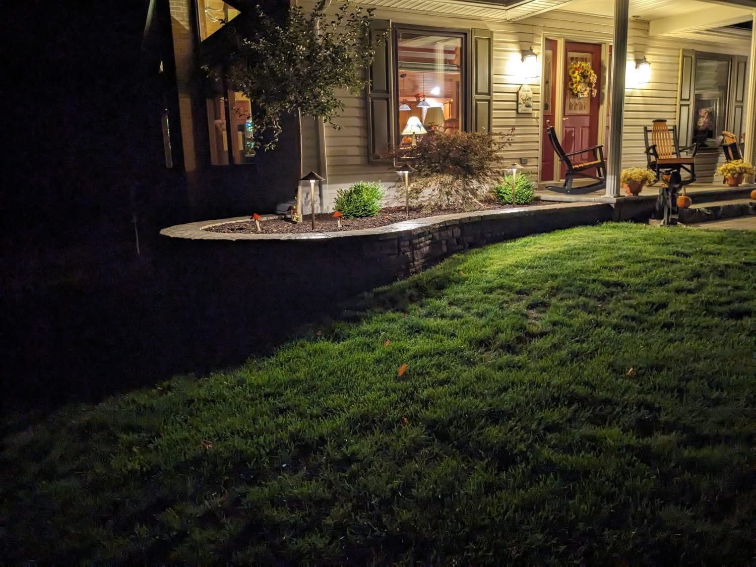 Night view of a house with lit porch, garden bed with a stone border, and a green lawn.