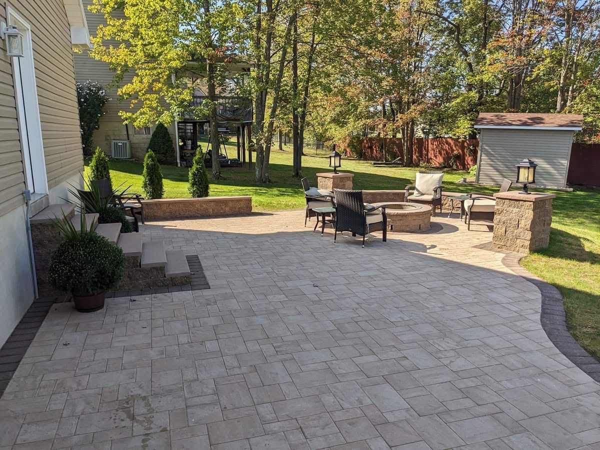 Brick patio with fire pit, seating, and landscaping next to a house and yard.