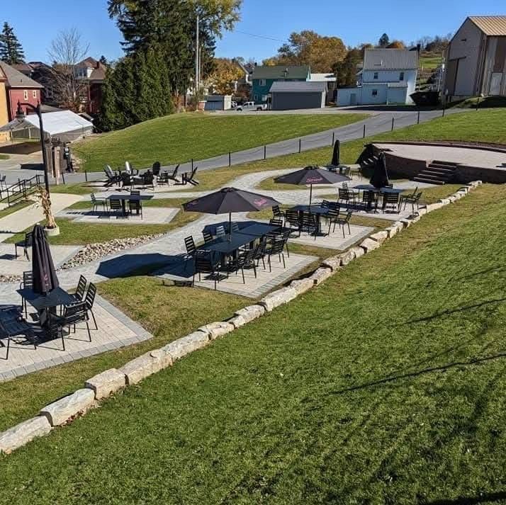 Outdoor patio with tables and umbrellas on a grassy hill; stone retaining wall, buildings in background.