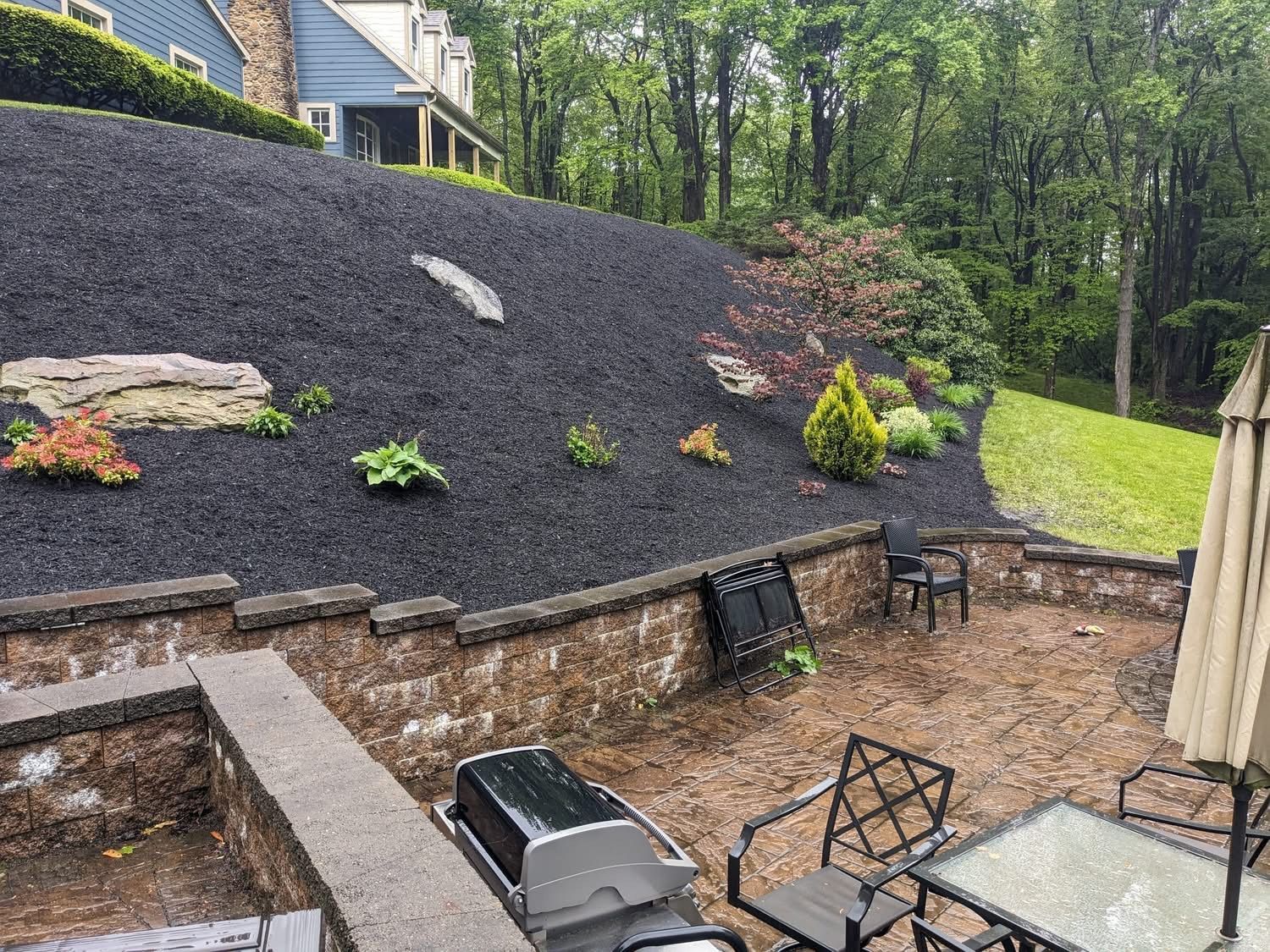 Patio with grill, table, chairs, and landscaped hillside with black mulch and plants.
