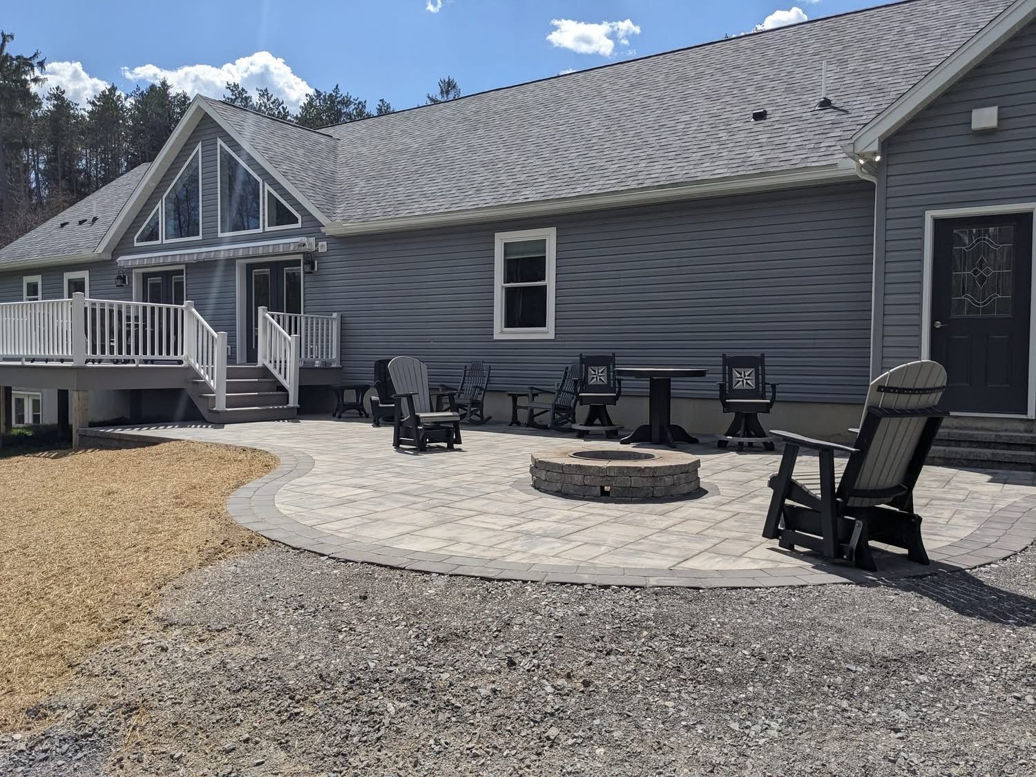 A house with a patio and fire pit. Several chairs surround the fire pit. Blue siding, a deck, and light gravel.