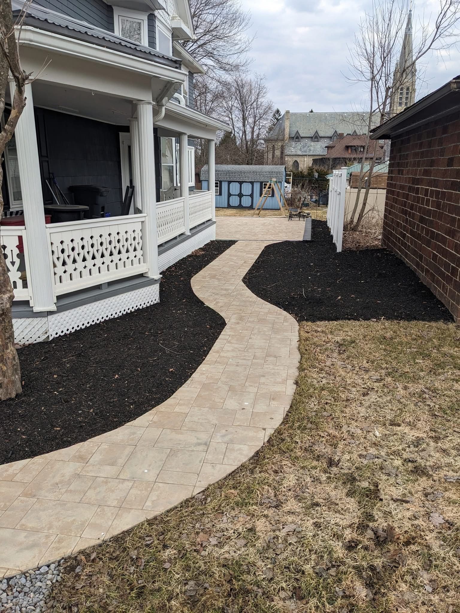 Stone walkway curves past a porch with white railing and a yard with black mulch and brown grass.