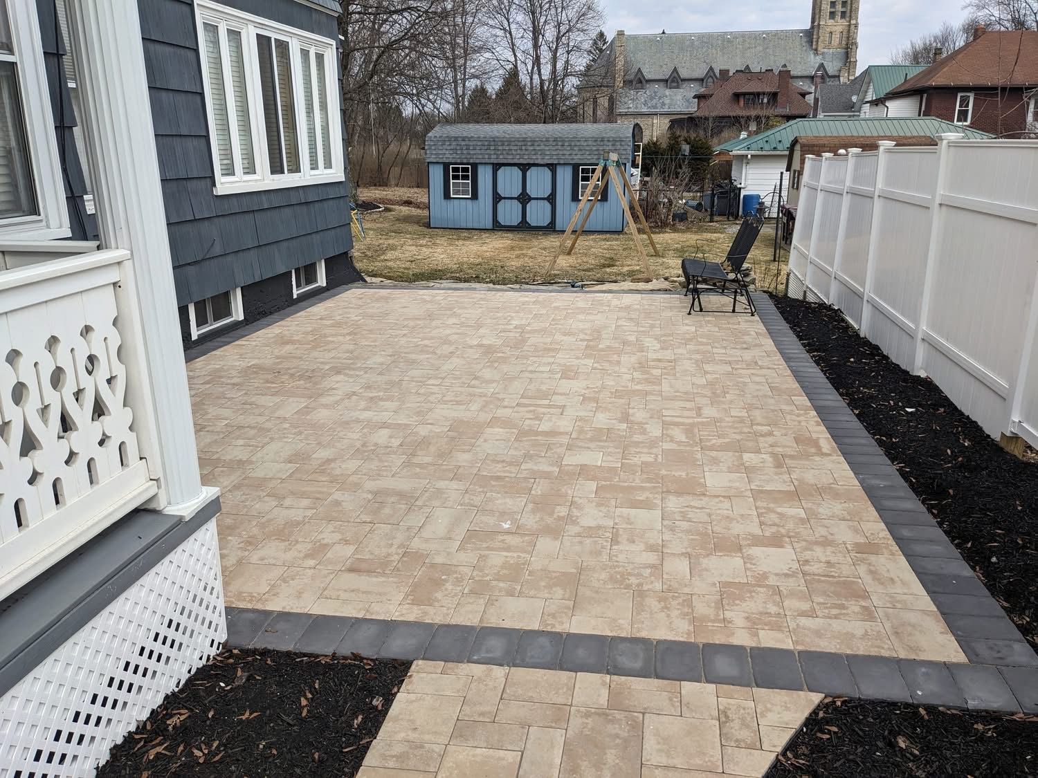 A paved patio with tan bricks, bordered by dark gray bricks and mulch. A blue shed and white fence are in the background.