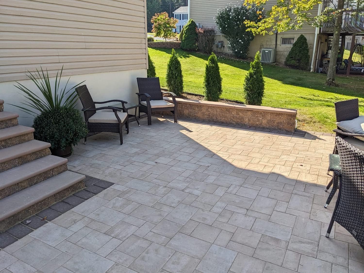 Patio with tan pavers, steps, seating, retaining wall with shrubs, and grassy yard.