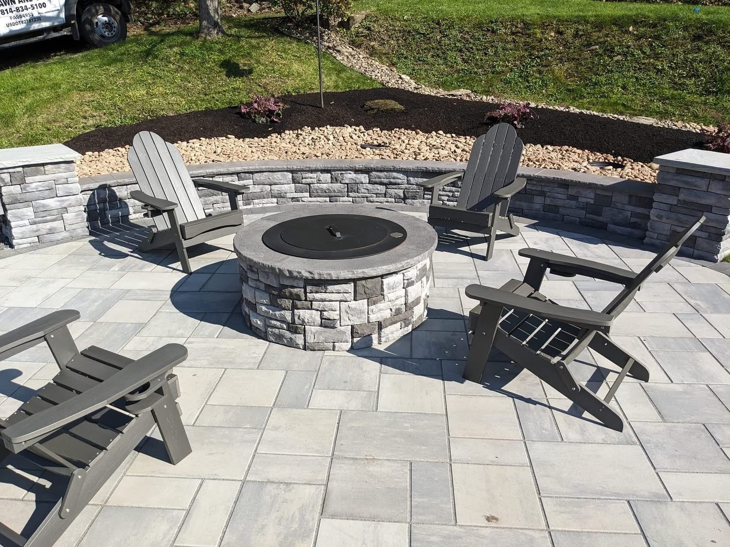 Stone fire pit with four Adirondack chairs on a paved patio, surrounded by a low stone wall and landscaping.