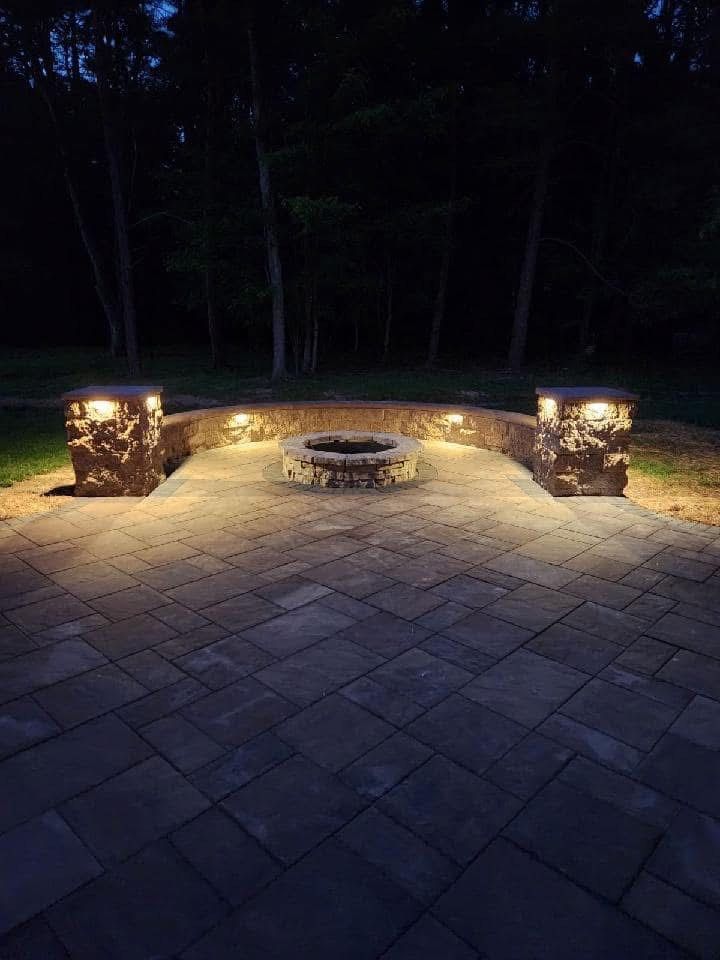 Lit outdoor patio with fire pit, stone wall, and trees at night.