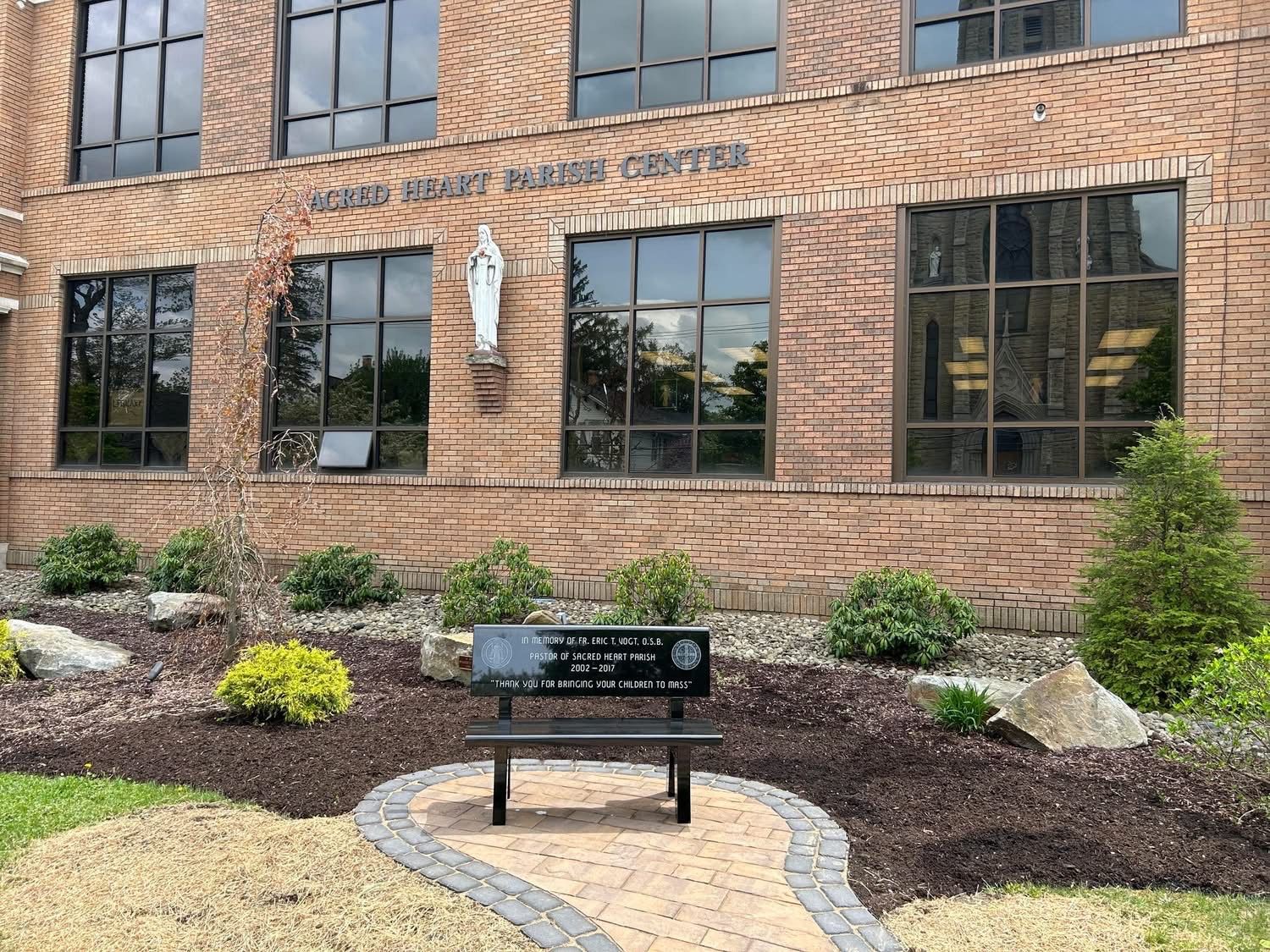 Brick building with windows. A bench sits in front with a plaque. Landscaping and a statue are visible.