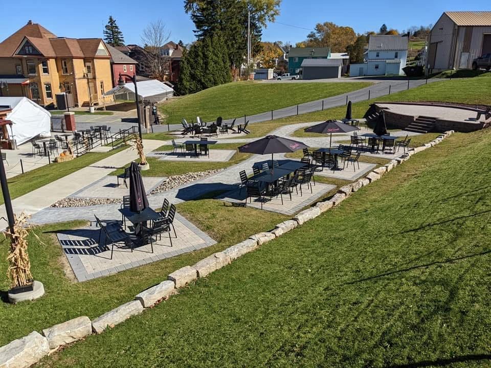 Outdoor seating area with tables and umbrellas on a grassy hillside. Buildings and trees in background.