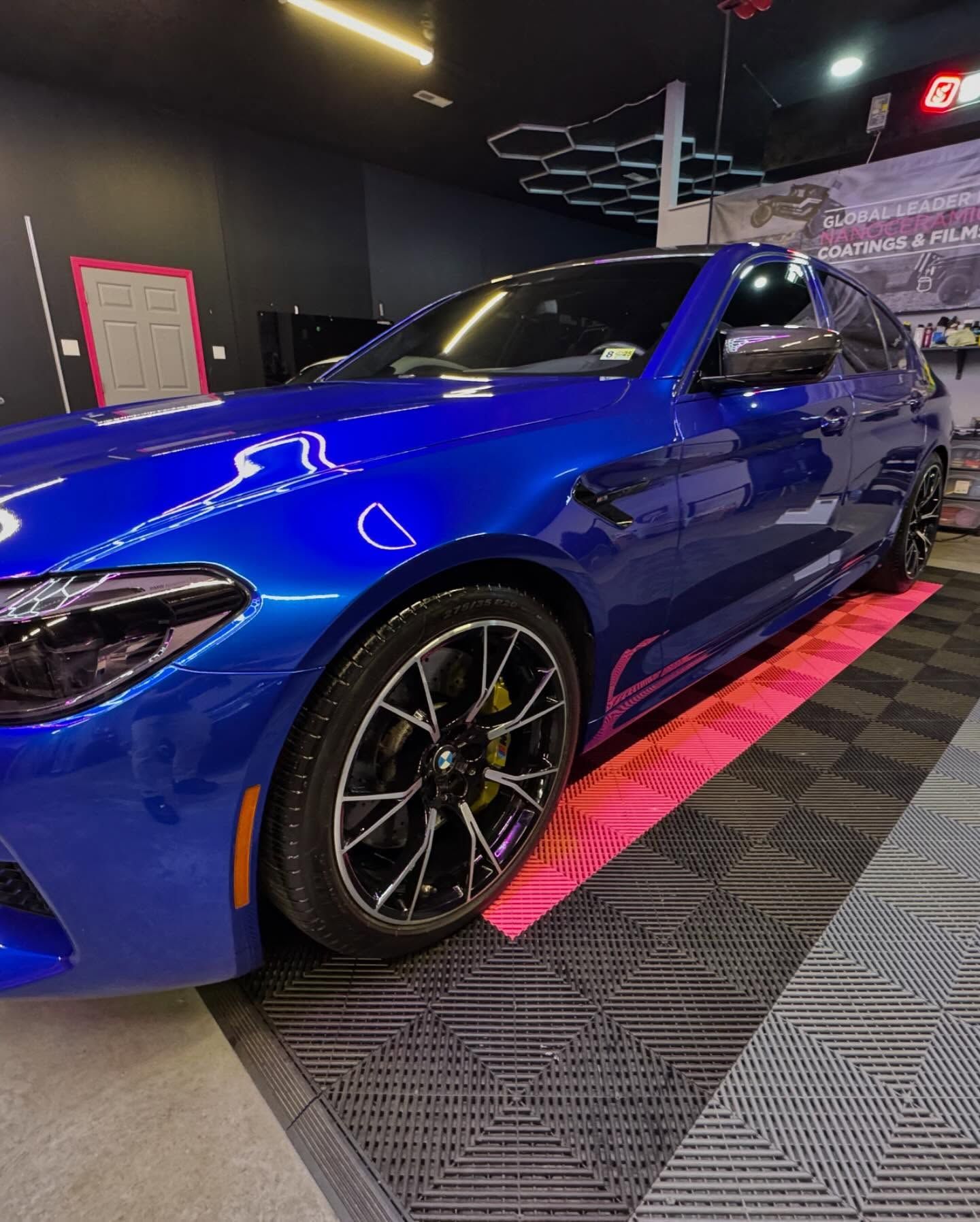 Blue BMW M5 car with black wheels parked in a garage with checkered flooring and pink mat.