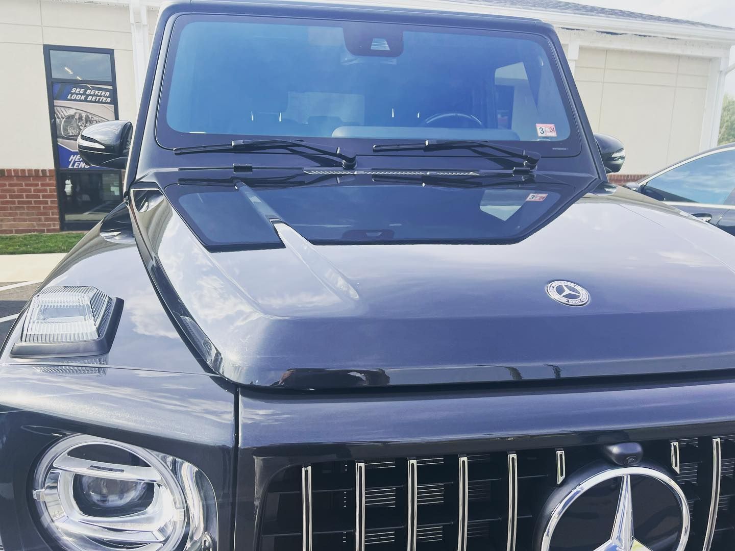Black Mercedes-Benz G-Class SUV, front view, parked outdoors, shiny finish, chrome grille, windshield reflection.