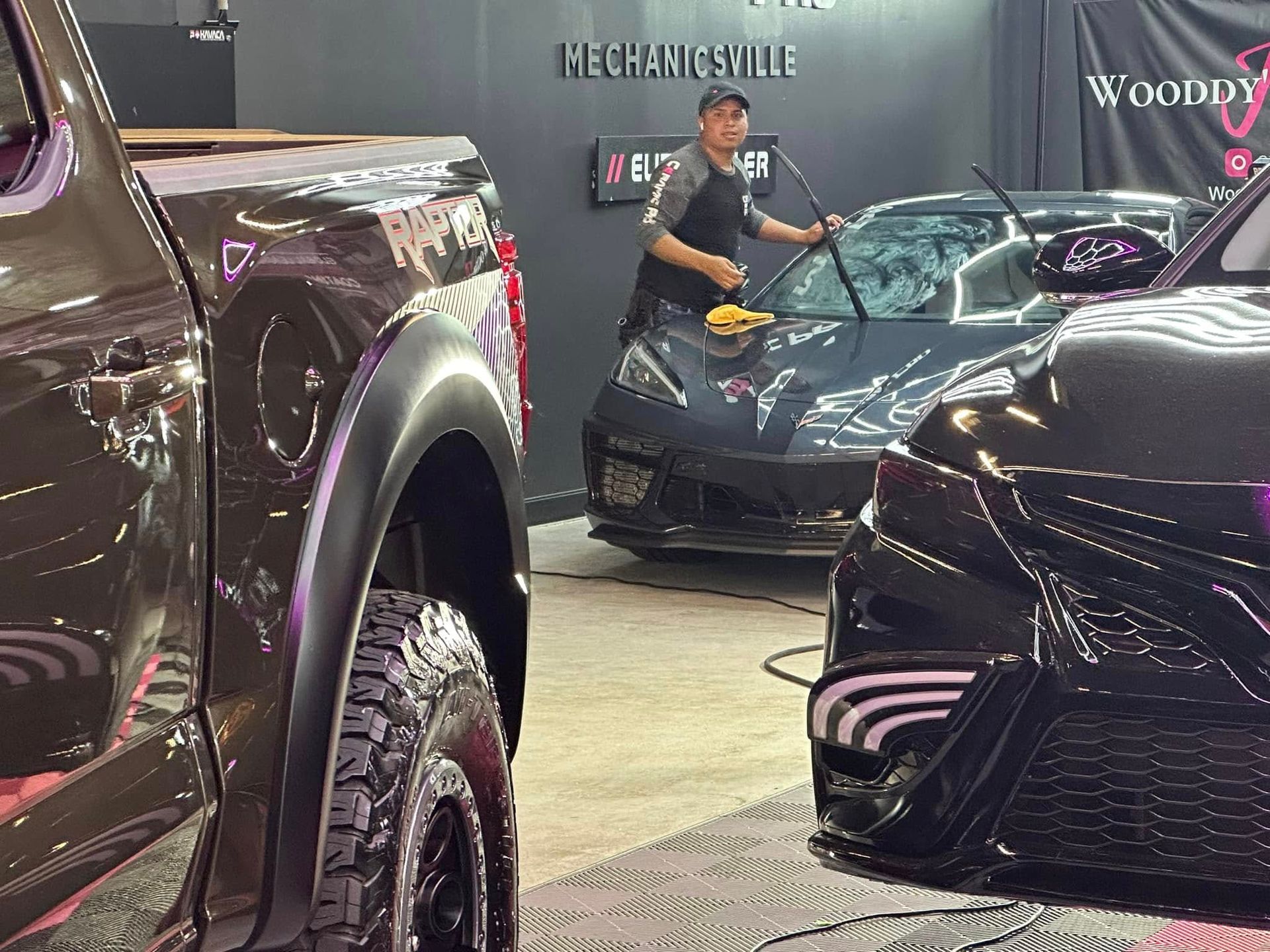 A person cleaning the windshield of a gray car inside a shop, with a brown truck and black car nearby.