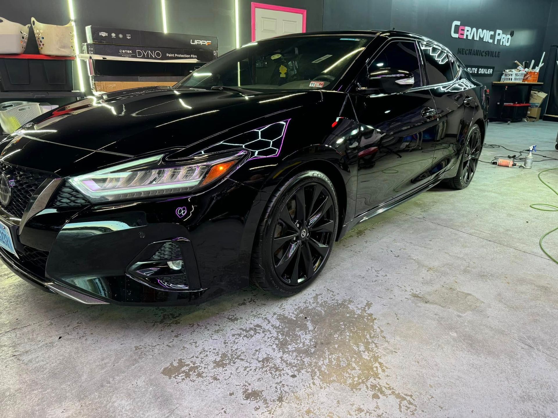 Black Nissan sedan in a detailing shop, glossy paint, black wheels.