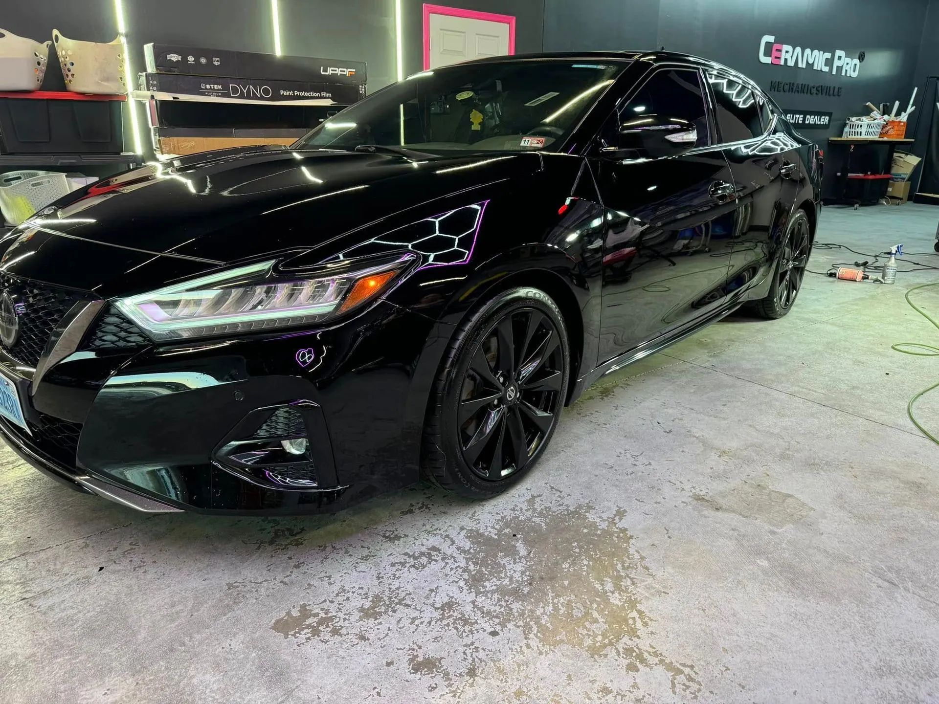 A black Nissan Maxima parked inside a Ceramic Pro auto detailing shop with glossy, reflective paint and black wheels.