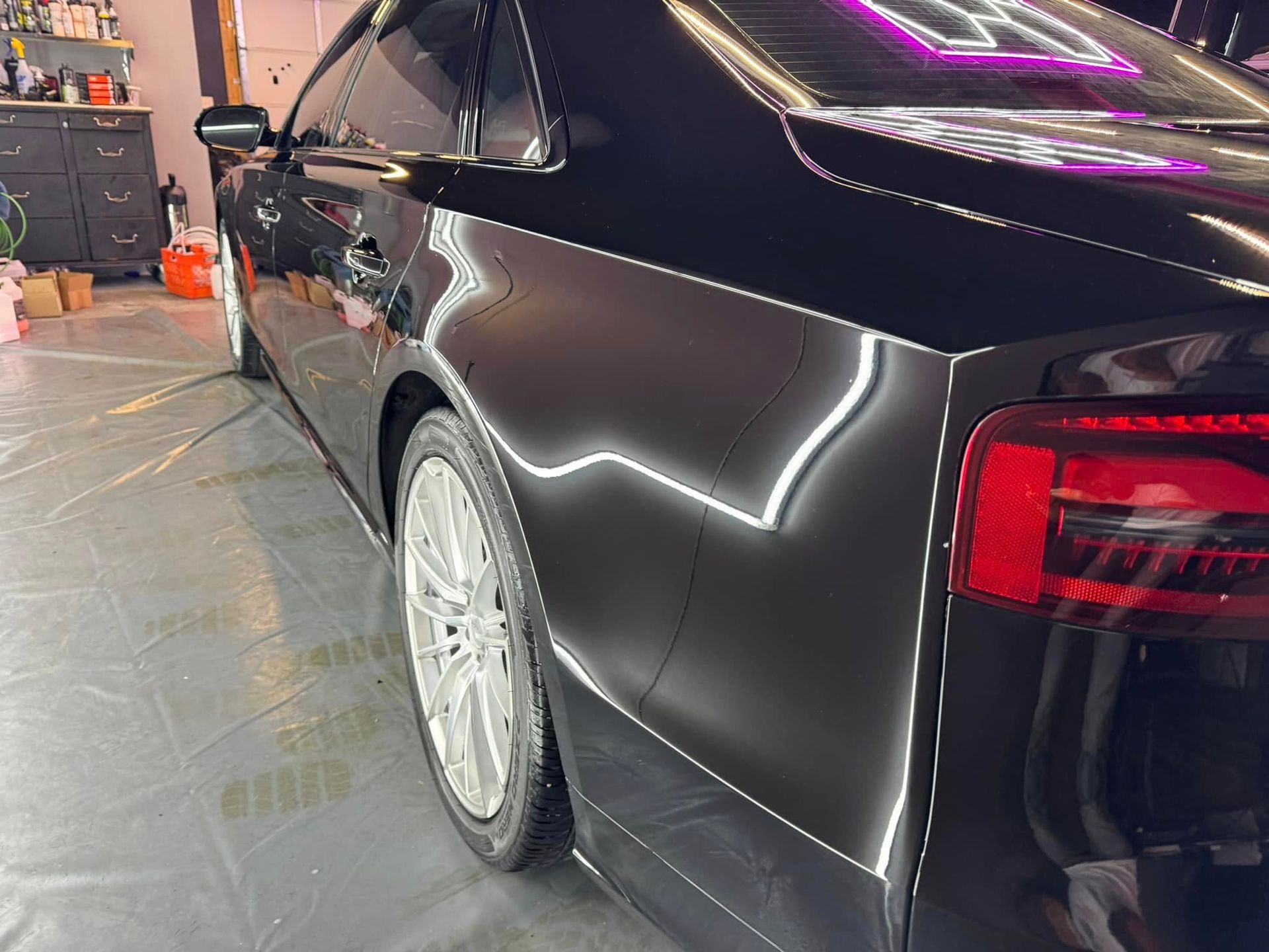 Black car's glossy finish reflects bright lights, highlighting the rear quarter panel and wheel.