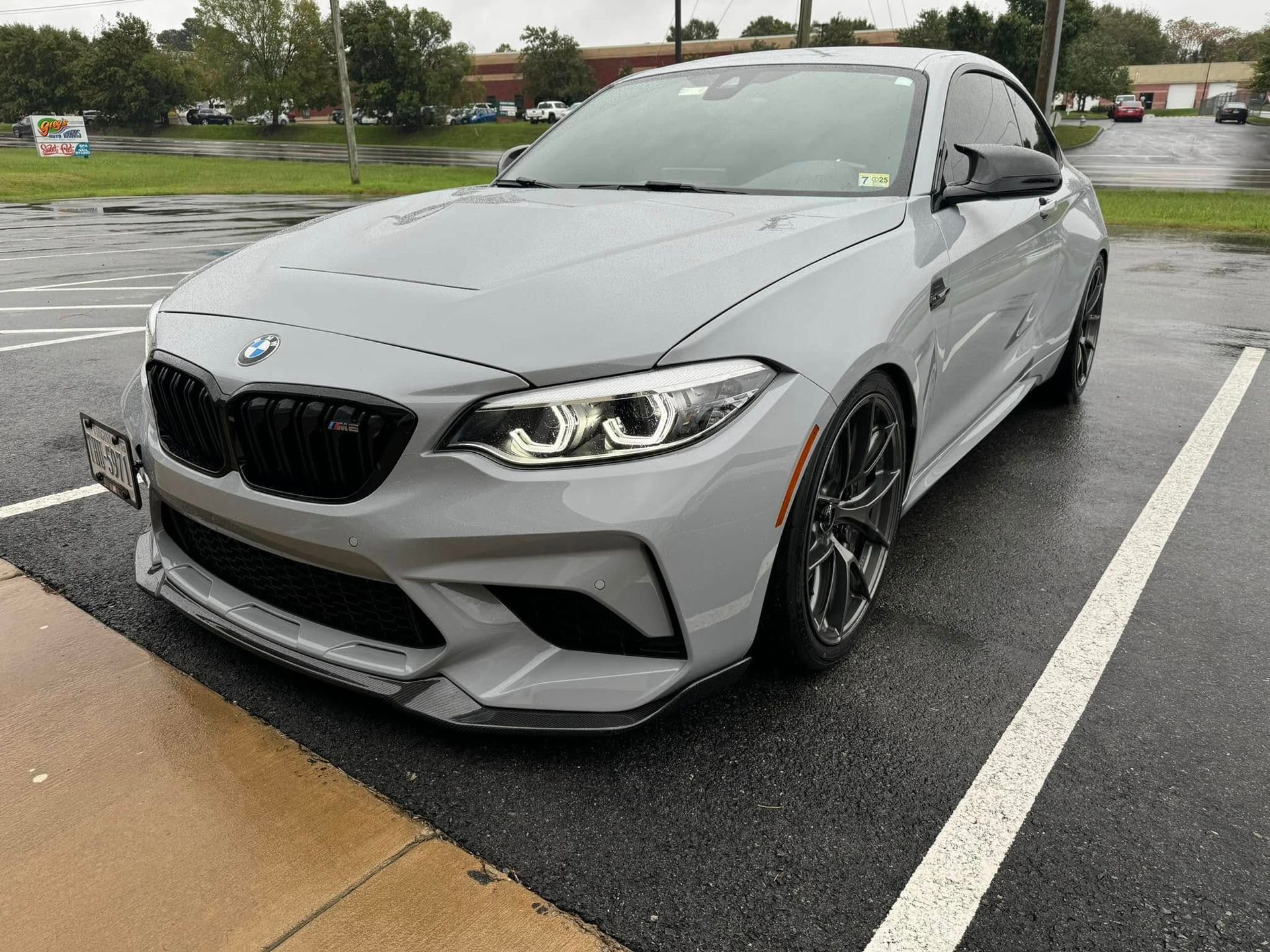 Gray BMW M2 coupe parked on wet asphalt, black grill and wheels.
