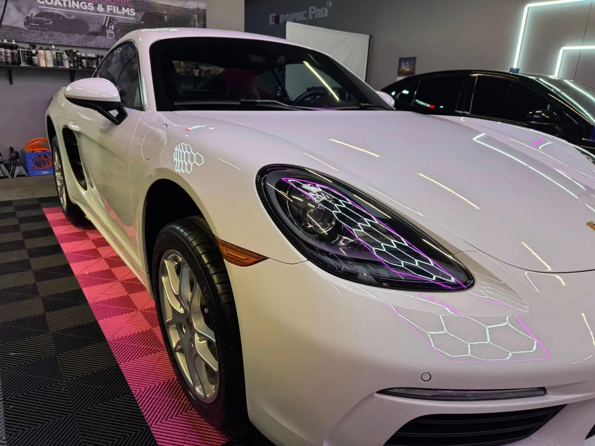A white Porsche parked indoors on a pink-and-black tiled floor, illuminated by overhead LED shop lighting.