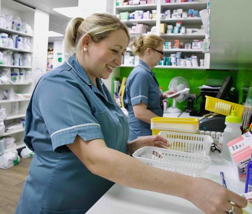 Two women are working in a pharmacy and one is holding a basket of pills.