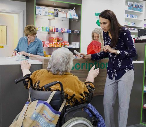 An elderly woman in a wheelchair is talking to a woman in a pharmacy.