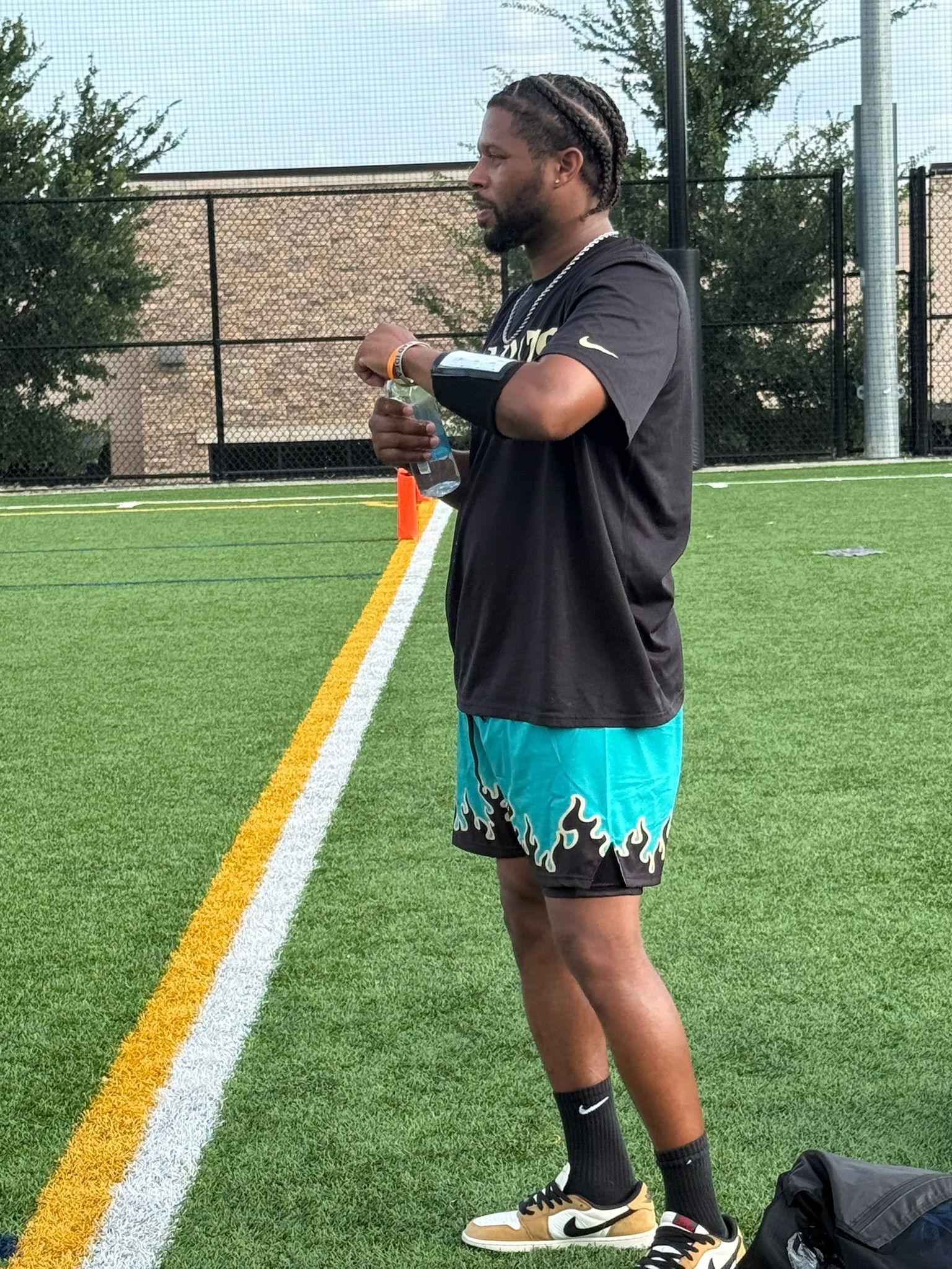 Man in teal shorts stands on a green sports field, holding a water bottle.