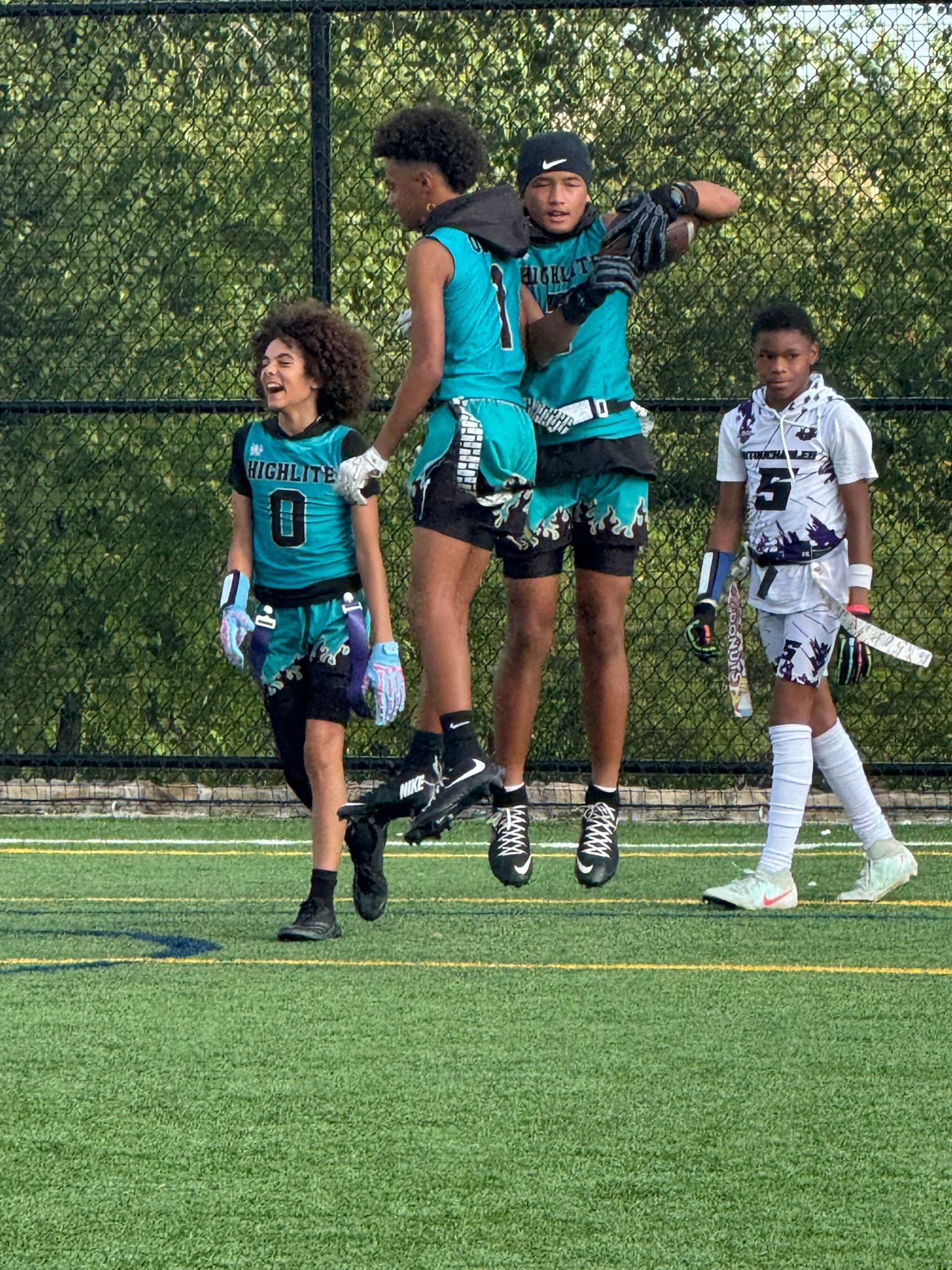 Flag football players in teal and white uniforms on a green field. One player jumps, celebrating.