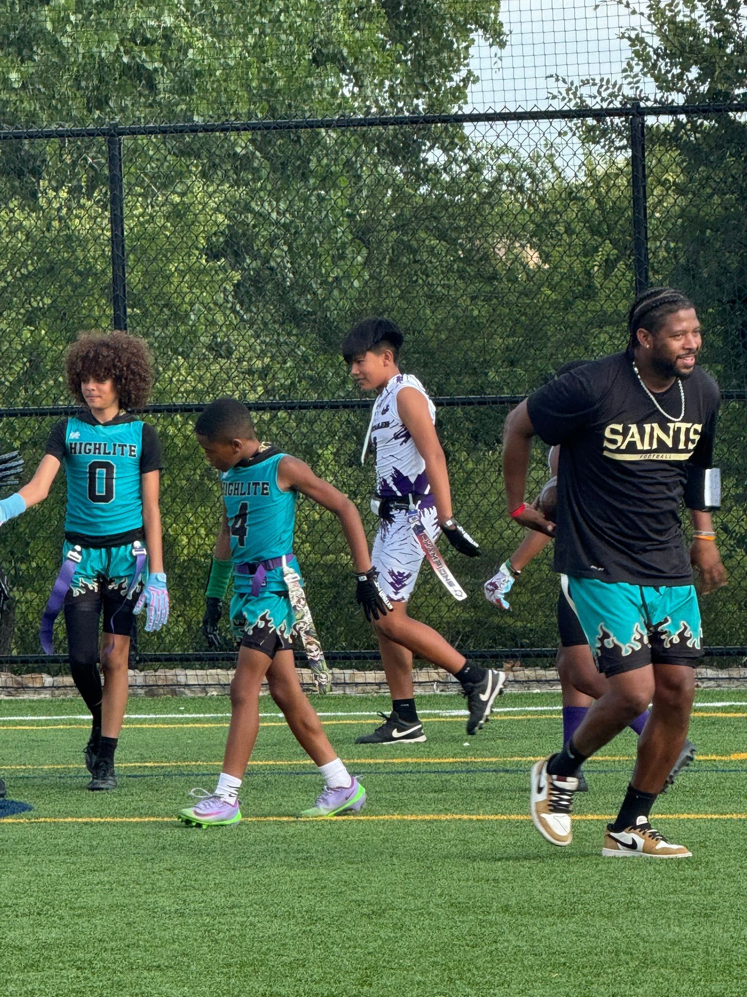 Flag football game: Youth players in teal and white jerseys, adult in Saints gear, on a green field.