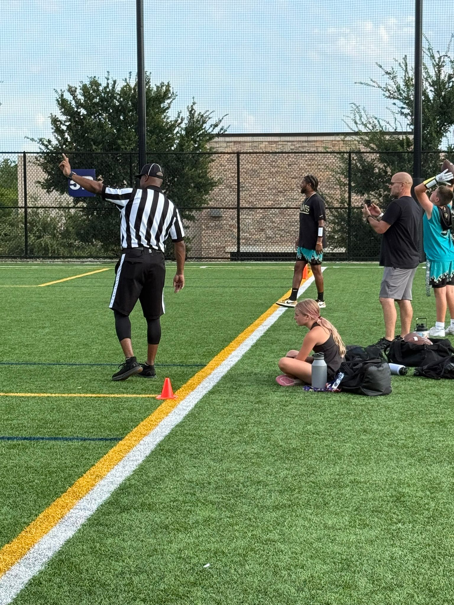 Referee signaling on a sports field, other players and a coach watch. Green grass, sunny.