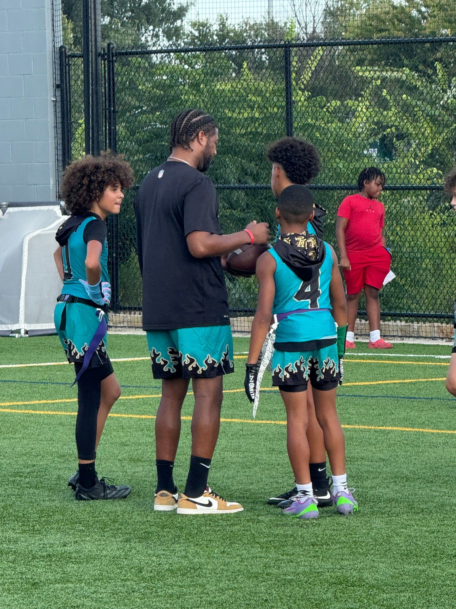 Group of people on a green field; a coach with kids in matching turquoise and black flag football attire.