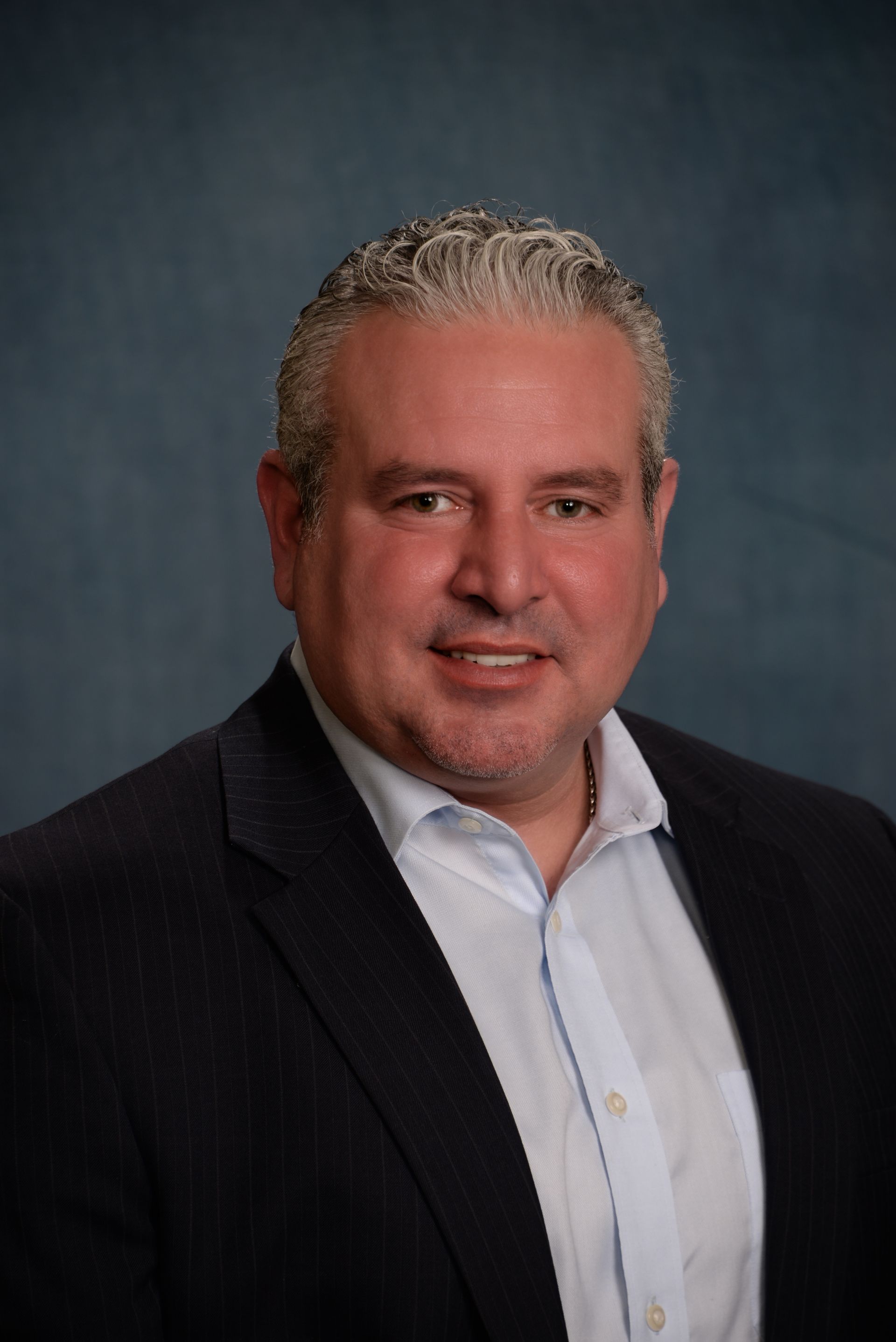 Man in a suit and open collar shirt smiles at the camera against a blue backdrop.
