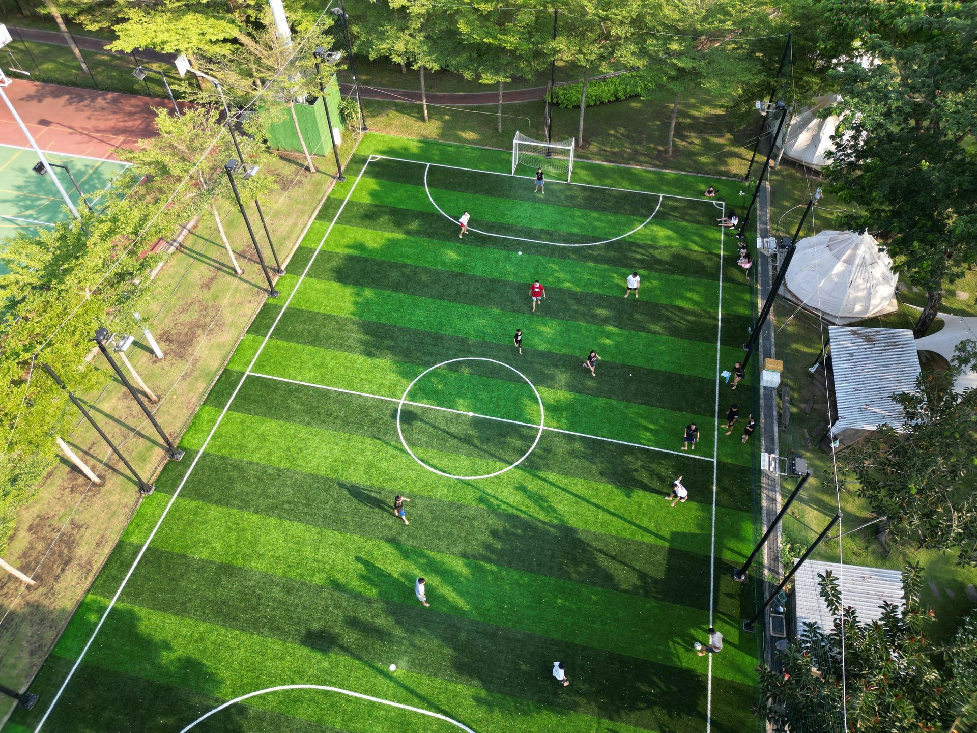 Aerial view of a soccer field with players in action, surrounded by trees and a tennis court.