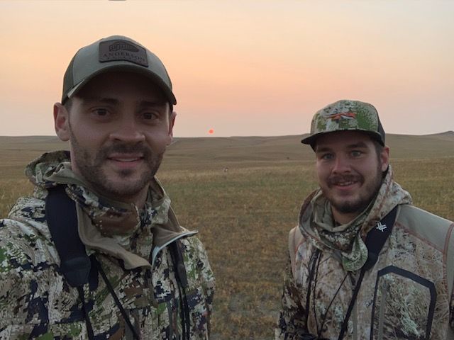 Two men are standing in a field with a sunset in the background.