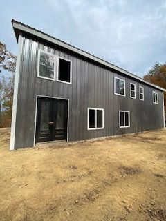 Gray metal-sided modern house with angled roof, multiple windows, and black front door set in a dirt lot.