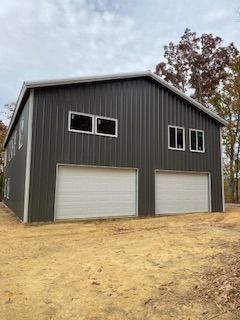 Two-story dark gray metal building with two garage doors and upper windows, set in a dirt yard with trees.